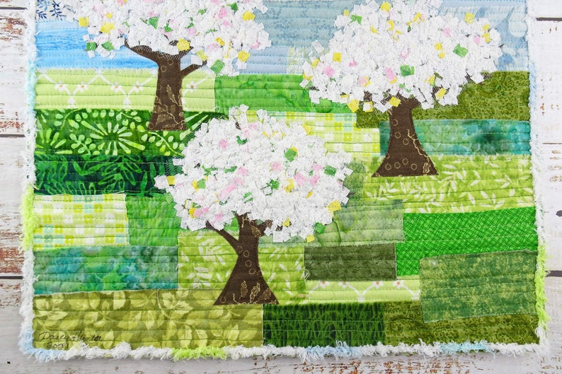 May include: A patchwork quilt featuring three stylized trees with white blossoms against a green and blue background.