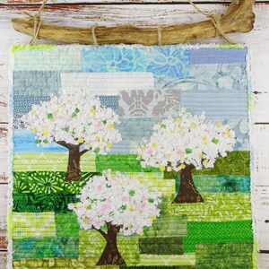 May include: A handmade wall hanging featuring a patchwork design of three trees with white blossoms against a green and blue background. The trees are made of fabric scraps and the background is a mix of different fabrics.