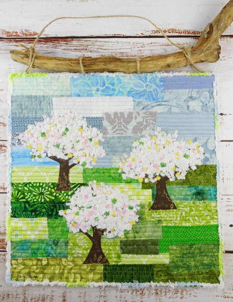 May include: A handmade wall hanging featuring a patchwork design of three trees with white blossoms against a green and blue background. The trees are made of fabric scraps and the background is a mix of different fabrics.