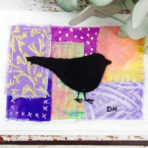 Peut inclure: Carte de vœux faite à la main avec une silhouette d'oiseau noir sur un fond patchwork. Le patchwork comprend des morceaux de tissu violets, roses et jaunes avec des coutures décoratives. La carte a une bordure blanche et les initiales "DH" sont visibles.