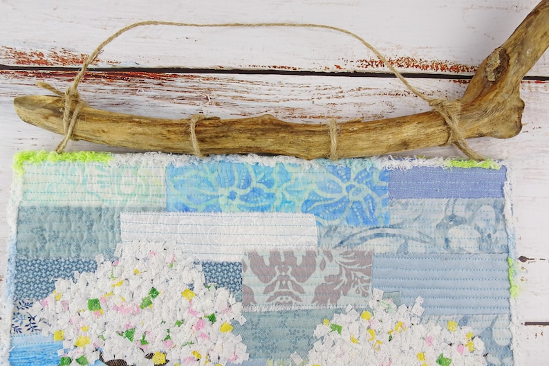 May include: A blue and white patchwork fabric wall hanging with a floral design. The hanging is attached to a natural branch with twine.