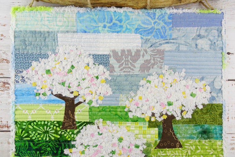 May include: A patchwork quilt wall hanging featuring two white flowering trees with brown trunks against a blue and green background.