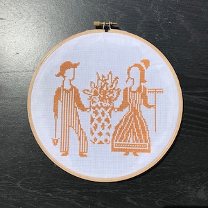 Op de afbeelding: Een witte borduurring met een bruin kruissteekontwerp van twee figuren die naast een boeket bloemen staan. De figuren dragen traditionele kleding en houden een boeket bloemen vast.