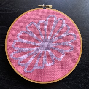May include: A pink and white cross-stitch embroidery of a stylized flower design in a wooden embroidery hoop.