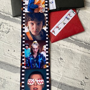 May include: A Stranger Things themed bookmark with four images of the character Eleven. The bookmark is designed to look like a film strip with numbers 15, 16, 17, and 18 along the side. The images show Eleven in different scenes from the show, including her wearing a blue shirt with a galaxy print, a blue jacket, and a pair of pizza-themed goggles.