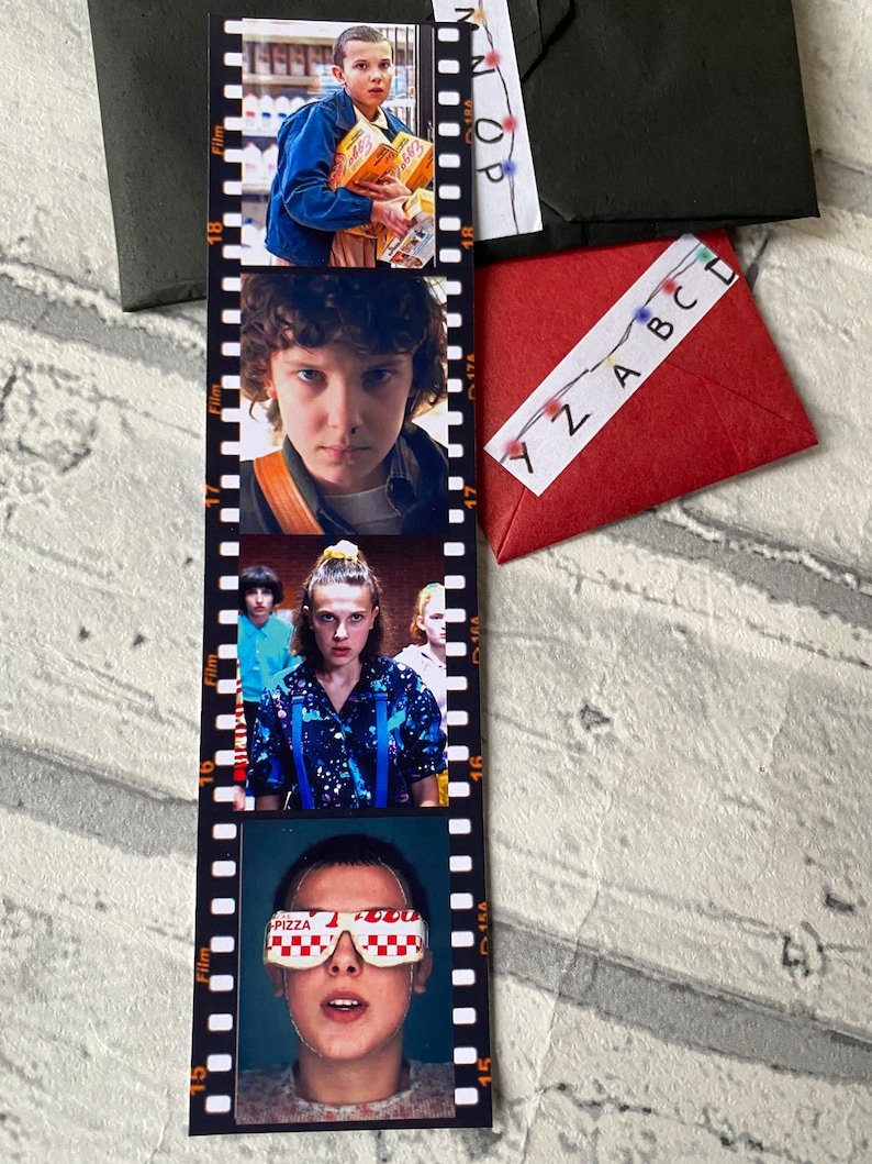 May include: A Stranger Things themed bookmark with four images of the character Eleven. The bookmark is designed to look like a film strip with numbers 15, 16, 17, and 18 along the side. The images show Eleven in different scenes from the show, including her wearing a blue shirt with a galaxy print, a blue jacket, and a pair of pizza-themed goggles.