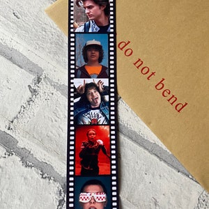 May include: A black and white film strip bookmark with five different images of characters from the show Stranger Things. The bookmark has the text "do not bend" printed on the right side.
