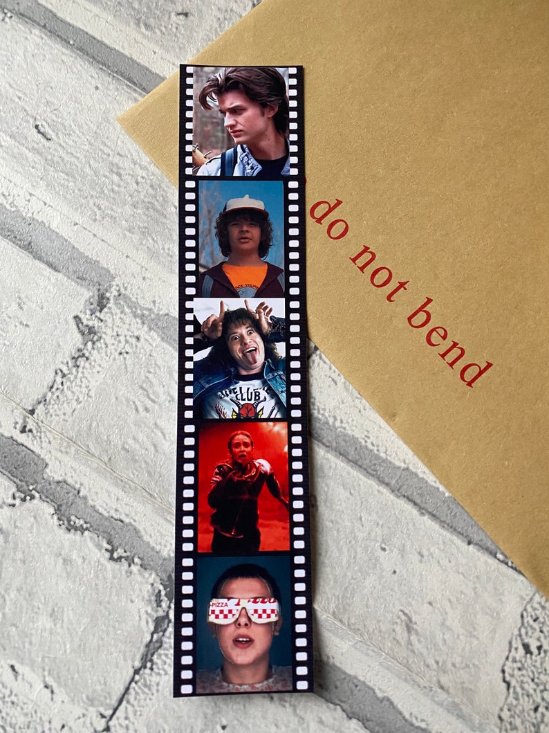 May include: A black and white film strip bookmark with five different images of characters from the show Stranger Things. The bookmark has the text "do not bend" printed on the right side.