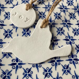 May include: Two white ceramic ornaments, one shaped like a bird and the other a circle with the word "JOY" embossed on it. Both ornaments are hanging from twine on a blue and white patterned background.