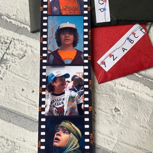 May include: A black and white film strip bookmark with four photos of a young boy wearing a baseball cap and different shirts. The photos are labeled with "Film" and "D15A", "D16A", "D17A", and "D18A". The text "HELLFIRE CLUB" is visible on one of the shirts.