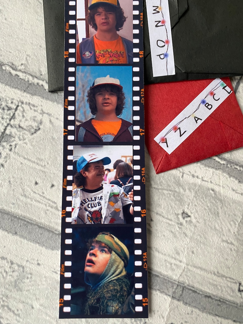 May include: A black and white film strip bookmark with four photos of a young boy wearing a baseball cap and different shirts. The photos are labeled with "Film" and "D15A", "D16A", "D17A", and "D18A". The text "HELLFIRE CLUB" is visible on one of the shirts.