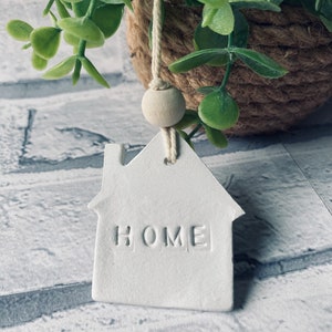 May include: White ceramic house-shaped ornament with the word "HOME" embossed on the front. The ornament is hanging from a white string with a wooden bead.