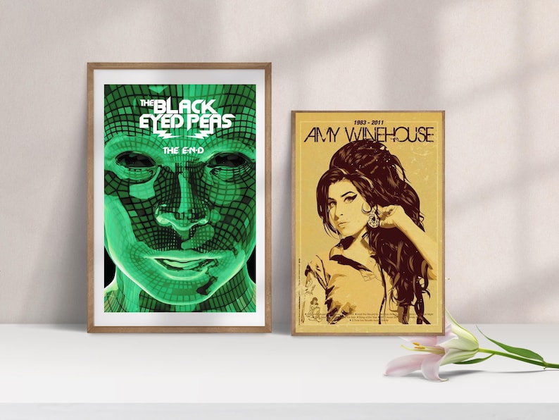May include: Two framed posters on a white surface. The left poster features a green face with the text "THE BLACK EYED PEAS THE E-N-D". The right poster is a portrait of Amy Winehouse with the text "1983-2011 AMY WINEHOUSE".