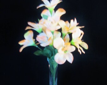 May include: A bouquet of artificial flowers with light orange and white petals featuring subtle dark speckles. The flowers are arranged in a tall, slender, clear glass vase with a metallic silver base, set against a solid black background.