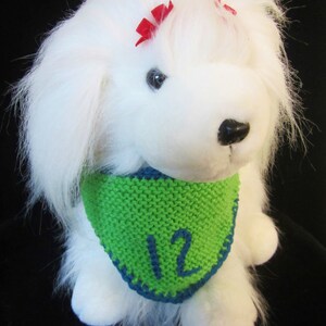 May include: A white plush dog with a green and blue knitted bandana. The bandana has the number 12 in blue. The dog has red bows in its fur and a black nose. The background is black.