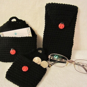 May include: A collection of handmade black crochet items, including a small purse, a glasses case, and a coin purse. Each item features a red ladybug button. A pair of eyeglasses rests nearby.