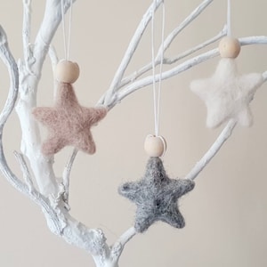 May include: Three felt star ornaments with wooden beads hanging from a white branch. The stars are in shades of gray, beige, and white.
