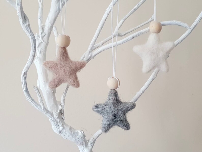 May include: Three felt star ornaments with wooden beads hanging from a white branch. The stars are in shades of gray, beige, and white.