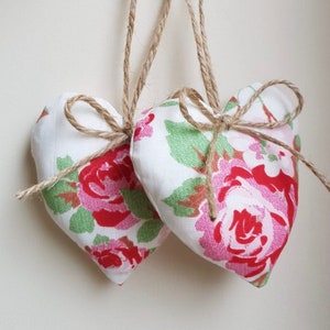 May include: Two white fabric heart-shaped ornaments with a floral pattern in shades of red, pink, and green. The hearts are tied with twine and have a rustic, farmhouse style.