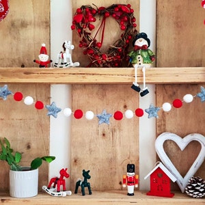 May include: A wooden shelf with a red and white felt ball garland with gray star accents. The shelf is decorated with Christmas ornaments, including a snowman, a rocking horse, a Santa Claus, and a nutcracker.