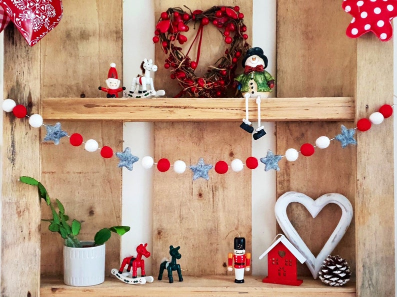 May include: A wooden shelf with a red and white felt ball garland with gray star accents. The shelf is decorated with Christmas ornaments, including a snowman, a rocking horse, a Santa Claus, and a nutcracker.