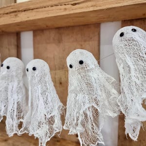 Op de afbeelding: Een slinger van witte spookdecoraties van gaas, elk met zwarte knoopogen. De spoken hangen voor een houten achtergrond, perfect voor Halloween of thema-evenementen.
