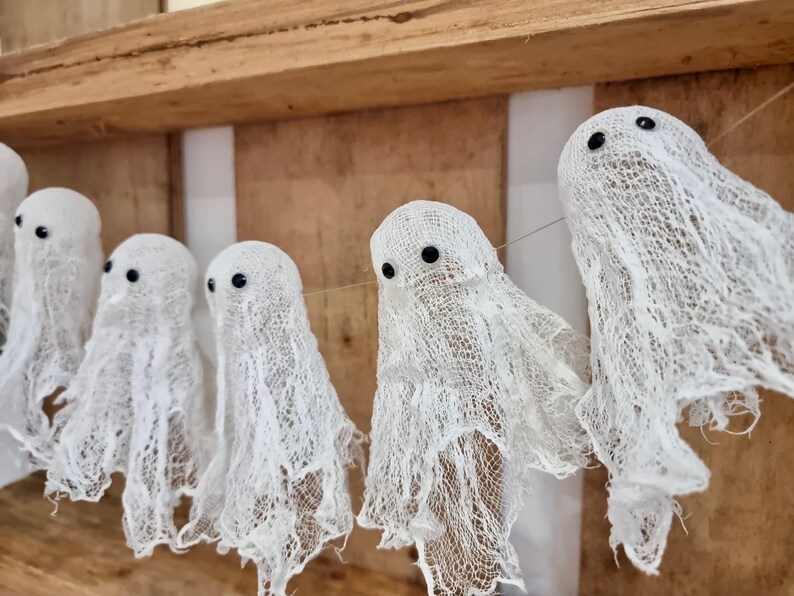 Op de afbeelding: Een slinger van witte spookdecoraties van gaas, elk met zwarte knoopogen. De spoken hangen voor een houten achtergrond, perfect voor Halloween of thema-evenementen.