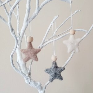 May include: Three felt star ornaments with wooden beads hanging from a white branch. The stars are in shades of beige, white, and gray.