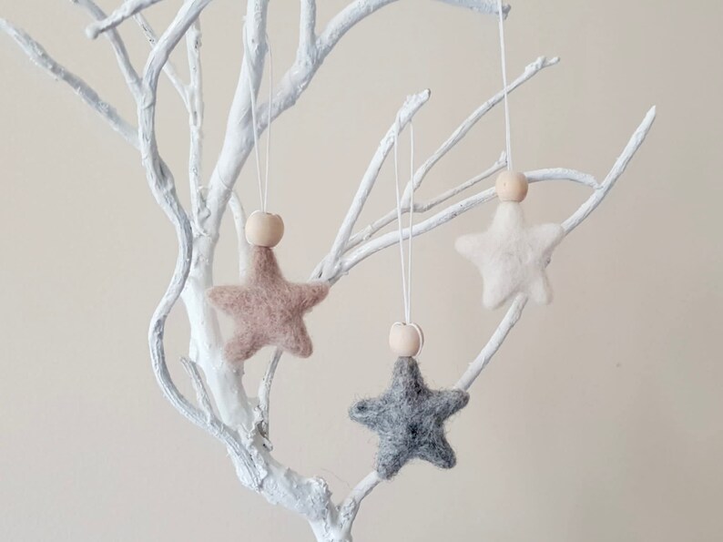 May include: Three felt star ornaments with wooden beads hanging from a white branch. The stars are in shades of beige, white, and gray.