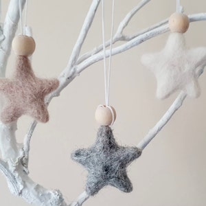 May include: Three felt star ornaments with wooden beads hanging from white branches. The stars are beige, white, and gray.