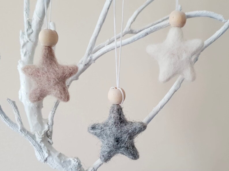 May include: Three felt star ornaments with wooden beads hanging from white branches. The stars are beige, white, and gray.