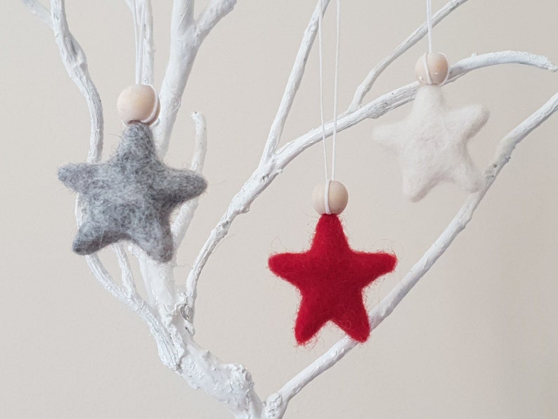 May include: Three felt star ornaments hanging from a white branch. The stars are gray, white, and red.