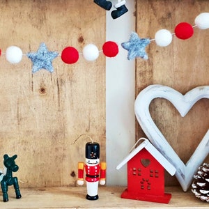 May include: A wooden shelf decorated with a red and white felt garland with gray stars, a white wooden heart, a red wooden house, a wooden nutcracker, and a pine cone.