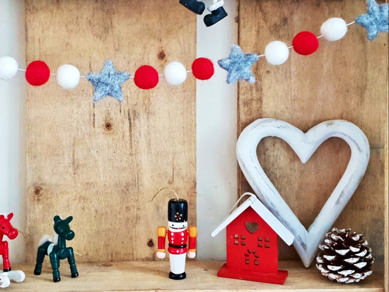 May include: A wooden shelf decorated with a red and white felt garland with gray stars, a white wooden heart, a red wooden house, a wooden nutcracker, and a pine cone.