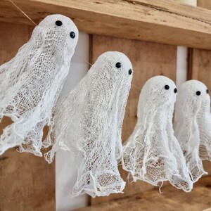 Op de afbeelding: Een slinger van witte spookdecoraties met zwarte ogen, gemaakt van een gaasachtig materiaal. De geesten hangen aan een touwtje tegen een houten achtergrond, perfect voor Halloween of spookachtige decoraties.