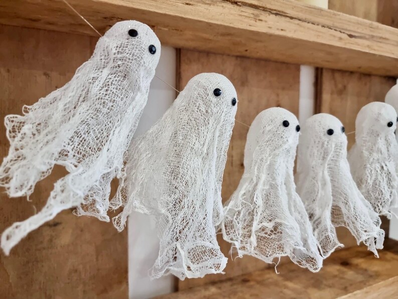 Op de afbeelding: Een slinger van witte spookdecoraties met zwarte ogen, gemaakt van een gaasachtig materiaal. De geesten hangen aan een touwtje tegen een houten achtergrond, perfect voor Halloween of spookachtige decoraties.