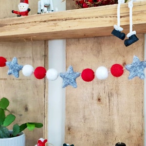 May include: A festive garland with white, red, and gray felt balls and stars. The garland is hanging on a wooden shelf.