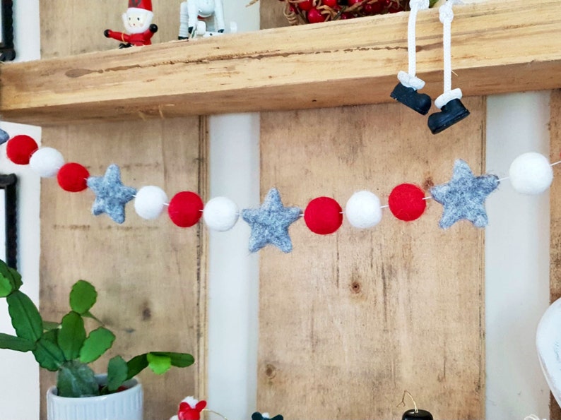 May include: A festive garland with white, red, and gray felt balls and stars. The garland is hanging on a wooden shelf.