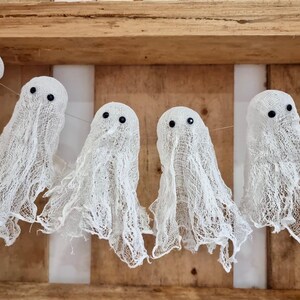 Op de afbeelding: Een slinger van witte spookdecoraties met zwarte kraaloogjes, tegen een houten achtergrond. De spoken zijn gemaakt van een gaasachtig materiaal, perfect voor Halloween.
