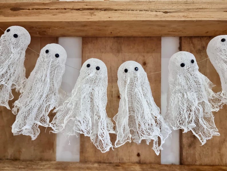 Op de afbeelding: Een slinger van witte spookdecoraties met zwarte kraaloogjes, tegen een houten achtergrond. De spoken zijn gemaakt van een gaasachtig materiaal, perfect voor Halloween.