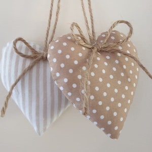 May include: Two heart-shaped decorations. One is beige with white polka dots, the other is beige with vertical white stripes. Both are tied with twine bows and have twine loops for hanging. Home decor.