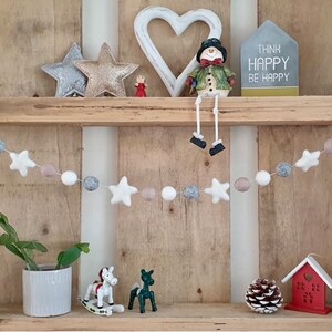 May include: A white, gray, and beige felt ball garland with white felt stars hanging on a wooden shelf. The garland is decorated with a snowman figurine, a heart-shaped sign, and a sign that reads "Think Happy Be Happy".