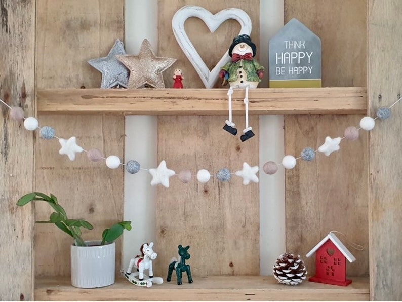 May include: A white, gray, and beige felt ball garland with white felt stars hanging on a wooden shelf. The garland is decorated with a snowman figurine, a heart-shaped sign, and a sign that reads "Think Happy Be Happy".