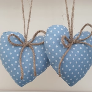 May include: Two heart-shaped decorations in pale blue with white polka dots. Each is adorned with a twine bow and suspended by twine. These fabric hearts are likely handmade, suitable for home decoration or as a gift.