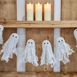 Op de afbeelding: Een Halloween-decoratie met een slinger van witte spookfiguren met zwarte ogen, opgehangen onder drie ivoorkleurige LED-kaarsen. De opstelling staat tegen een houten achtergrond en cre&euml;ert een spookachtige sfeer.