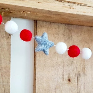 May include: A red, white, and gray felt garland with stars and pom-poms. The garland is strung on a white string.