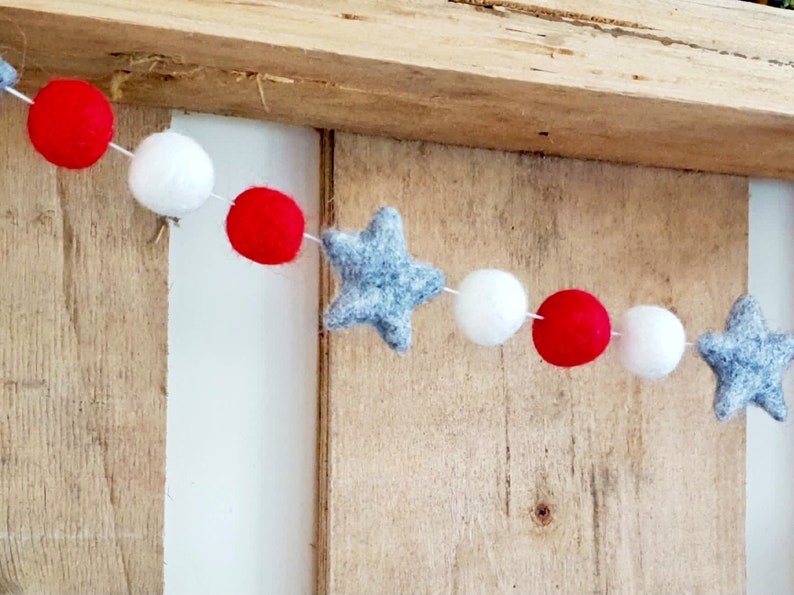 May include: A red, white, and gray felt garland with stars and pom-poms. The garland is strung on a white string.