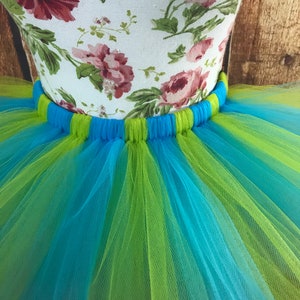 May include: A close-up of a colorful tutu skirt. The skirt is made of layers of blue and lime green tulle. The waistband is blue and green. The mannequin form is covered in a floral fabric with pink roses.