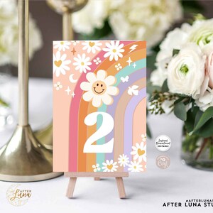 May include: A colorful table number card with a retro design. The card features a rainbow, daisies, and the number 2. Text at the top reads "INSTANT DOWNLOAD EDITABLE TEMPLATE". The card is displayed on a small wooden easel.
