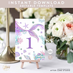 May include: Purple mermaid tail table number card with a white background. The card features a purple mermaid tail with iridescent scales, seashells, starfish, and coral. The number "1" is in the center of the card in a purple font. The card is on a small wooden easel.